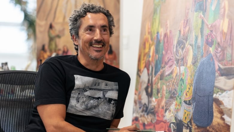 Photo of the artist Kent Monkman in his studio. He is a slim middle-aged man with short, wavy salt-and-pepper hair and a light beard. He smiles and wears a black graphic tee. He is seated in front of a large painted canvas, a paintbrush in his hand.