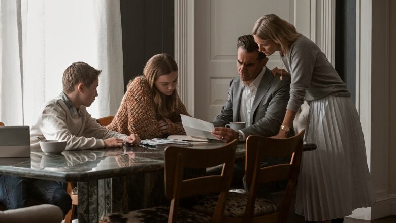 A family sits at a dinner table, all intently looking at a letter. Seated at the table, from left, is a young boy, a teenage girl, a man and a woman. The man is holding the letter, while the woman stands over him with her hand on his shoulder. 