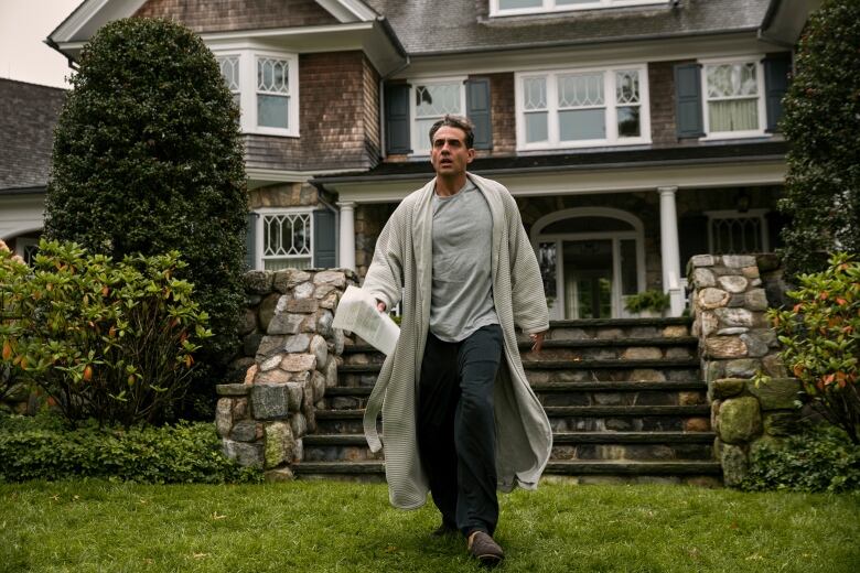 A man in a robe, slippers and pajamas strides out of a large home, holding a letter in his hand. He is walking on the grass toward the camera and appears stressed. 