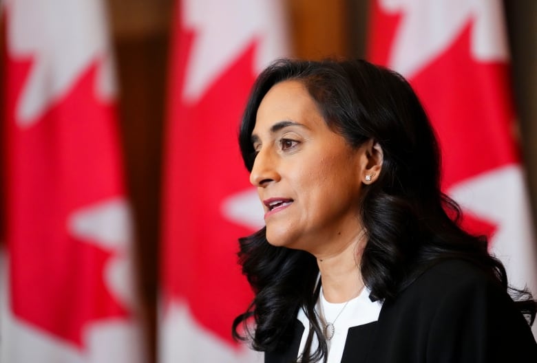 A woman with long black hair, pictured in profile, her mouth open mid-sentence, against a Canadian flag backdrop. 