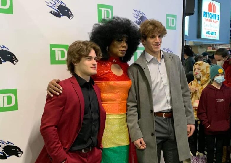 Three individuals, two white and one Black, stand in front of a marquee while their photo is being taken at a public event. The Black man is in drag and is wearing a dress made of the Pride rainbow colours. 