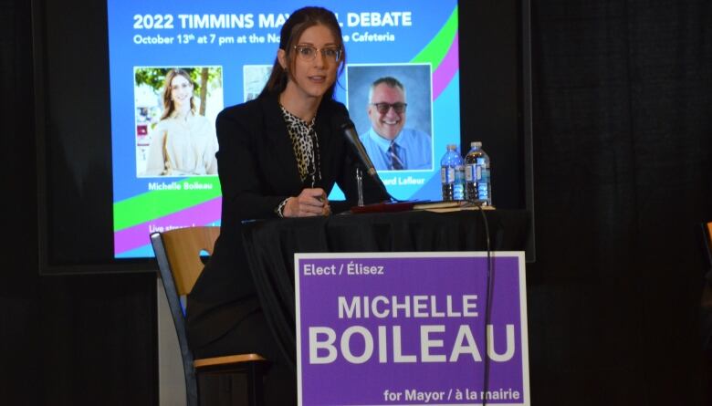 Michelle Boileau sits at a podium, with a purple sign bearing her name on the front