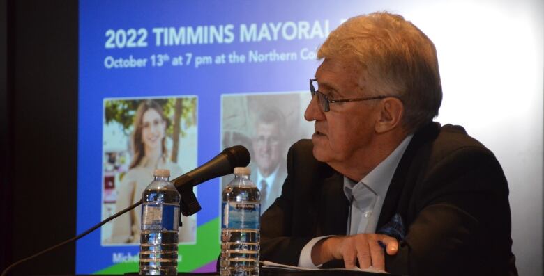 Joe Campbell sits at a podium, with two bottles of water.