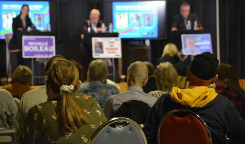 People sit in chairs and listen to what three candidates up on stage are saying