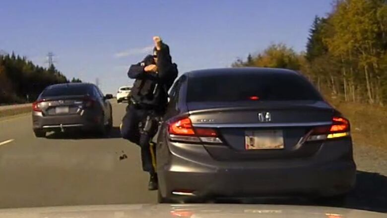 An RCMP officer shrinks away from the road, next to his vehicle, as a car passes him on a highway.