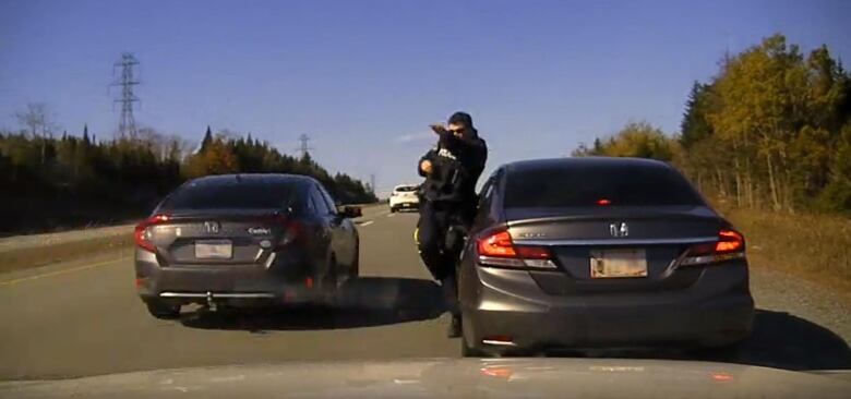 An RCMP officer shrinks away from the road, next to his vehicle, as a car passes him on a highway.