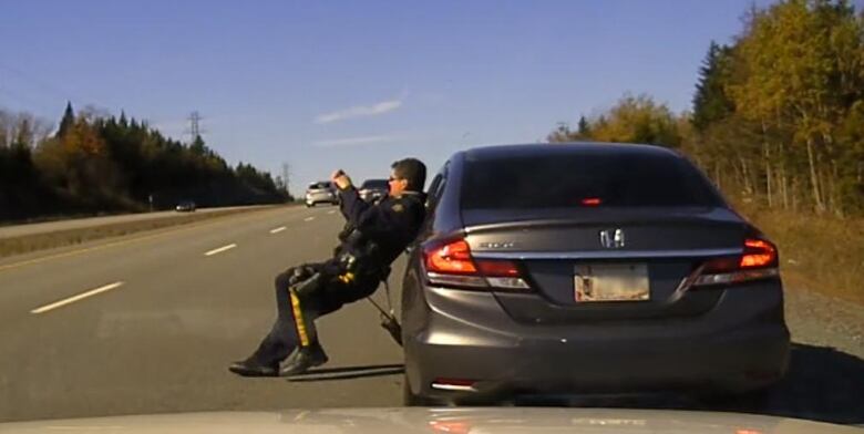 An RCMP officer shrinks away from the road, next to his vehicle, as a car passes him on a highway.