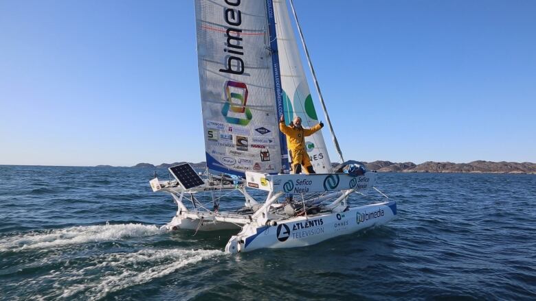 A man in a yellow onesie holds his hands in the air triumphantly aboard a catamaran as he sails out into the water.