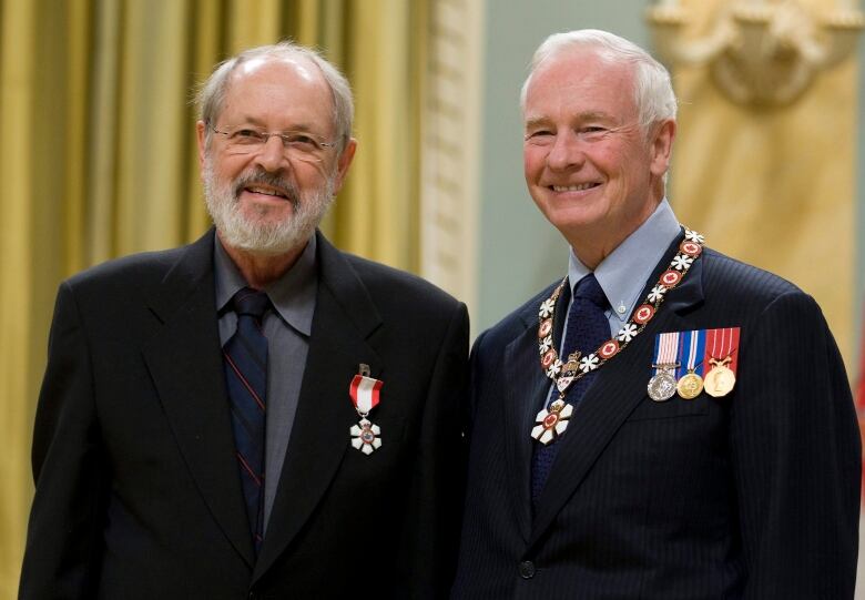 A bearded man in a suit jacket, adorned with a red-and-white Order of Canada medal on the lapel, smiles as he stands beside another man.