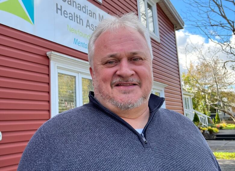 A middle-aged man smiles into the camera. The sign behind him says 