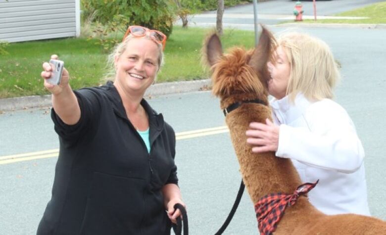 A woman holding an alpacas leash takes a picture of another woman posing with the animal.