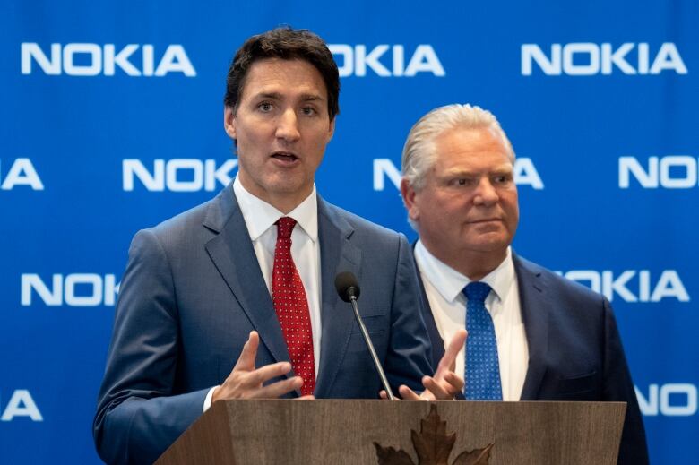 A politician speaks at a lectern in front of a blue corporate backdrop while another listens behind him.