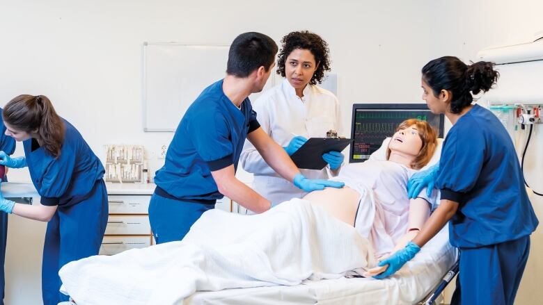 On the left, two women in blue scrubs both hold a medical device, wrapped in fuchsia cloth. On the right, a doctor in a white coat holds a clipboard. A woman and a man surround a mannequin on the hospital bed. The man has his gloved hand on the mannequin's stomach and the woman holds the mannequin's shoulder and hand. 