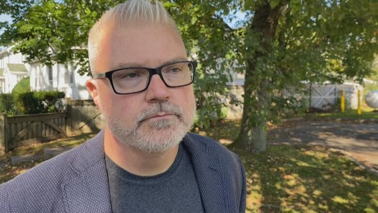 A man is seen with grey hair slick back from his facing and wearing black rectangular glasses, a navy blazer with white polka dots and a dark grey t-shirt.