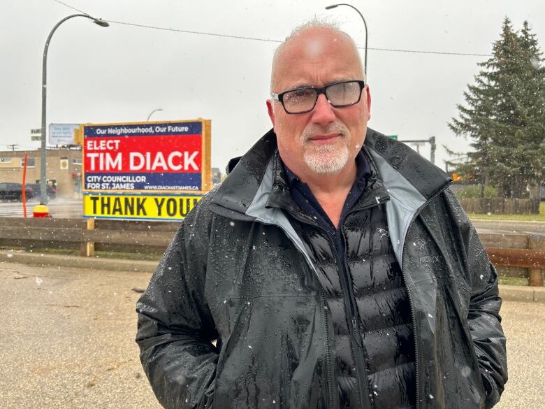 A man wearing glasses and a black jacket is standing in front of a sign that says 