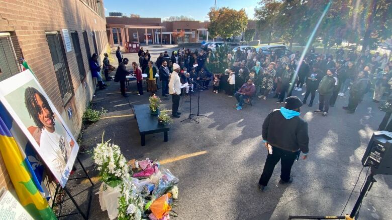 a crowd gathers around a large picture of a Black teen who was killed. flowers and candles are laid on the ground