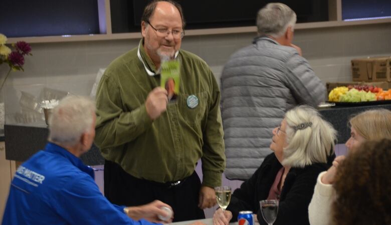 Chris Mayne stands next to a table, holding up a nametag around his neck, while Jane  Labb smiles, a glass of wine in front of her.