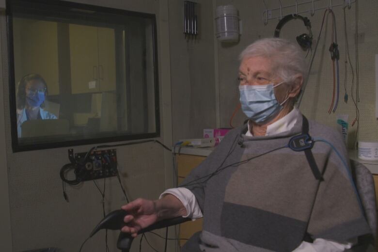 A silver-haired woman wearing a mask sits in a room as an audiologist watches from a nearby window.