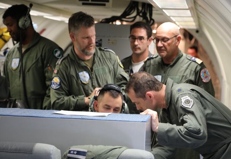 Six men in military uniforms sit and stand in front of a radar screen on an airplane. 