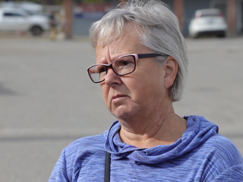 A woman short grey hair and glasses stands in a hospital parking lot. 