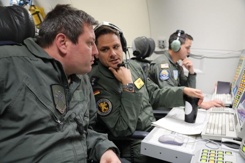 Three men wearing military uniforms and headsets sit in front of radar screens and keyboards on an AWACS aircraft.  