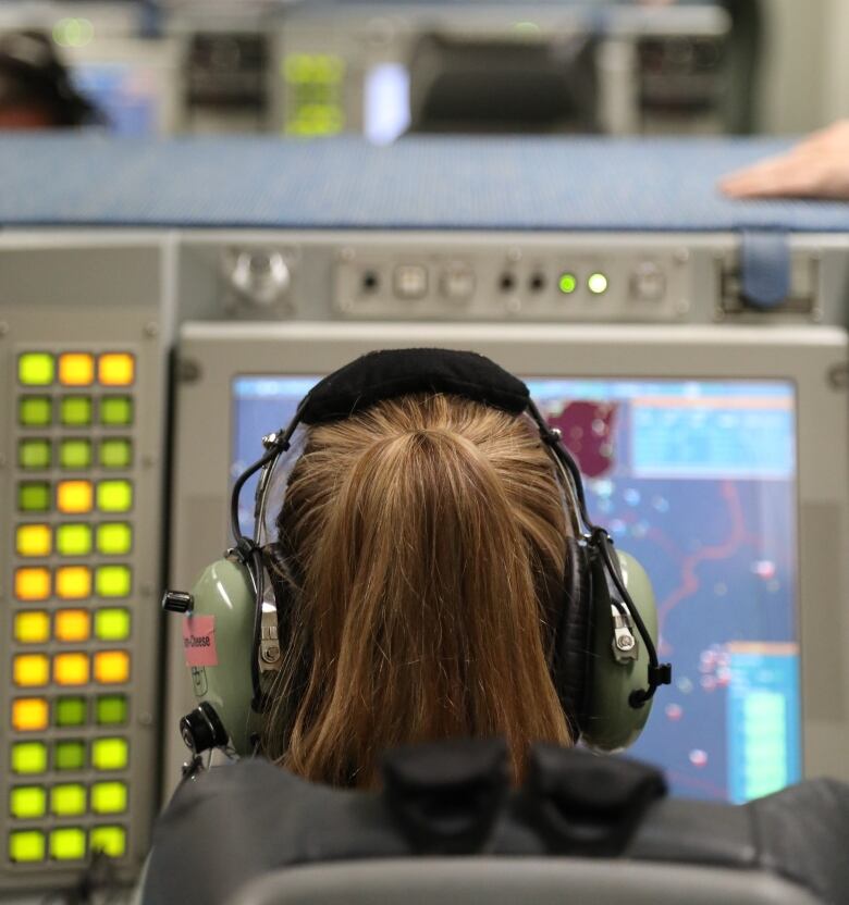 The back of a woman's head is seen as she wears headphones and watches a radar screen aboard an AWACS aircraft. 