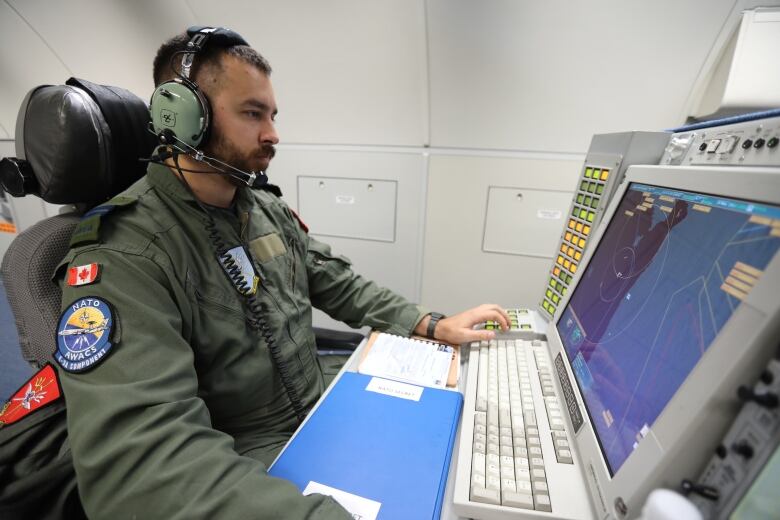 A man in a military uniform with a Canadian flag patch on his right shoulder wears headphones and watches a radar screen on an AWACS aircraft. 