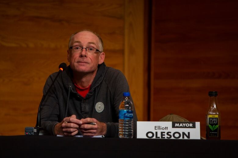 A bald man sits at a table smiling with mic in front of him.