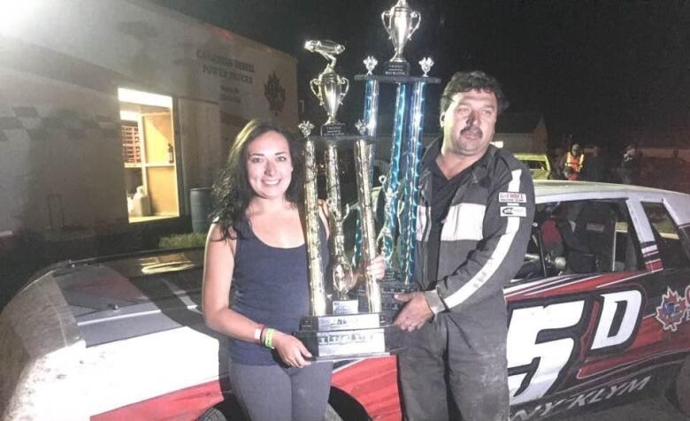 A young woman and middle-aged man hold large trophies in front of a race car.