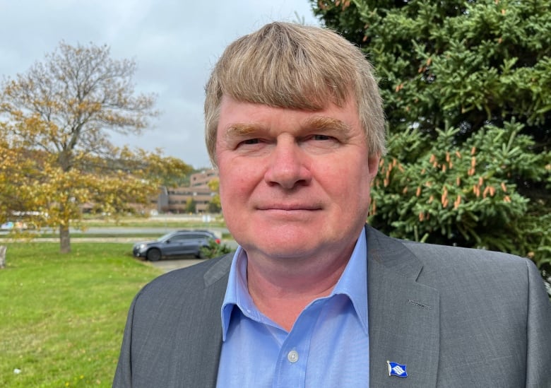 A man with greying hair and wearing a blue dress shirt and grey blazer stands outdoors with an evergreen tree and green grass in the background.