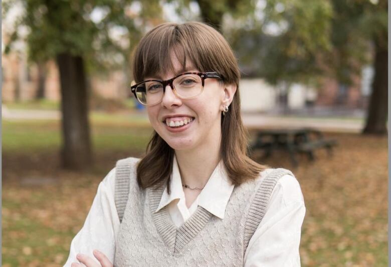 Person with brown shoulder-length hair and glasses, wearing dress shirt and vest, stands outside with fall colours in background.