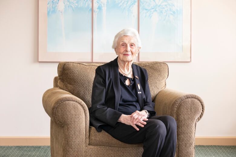 An older woman poses, sitting in a living room chair.