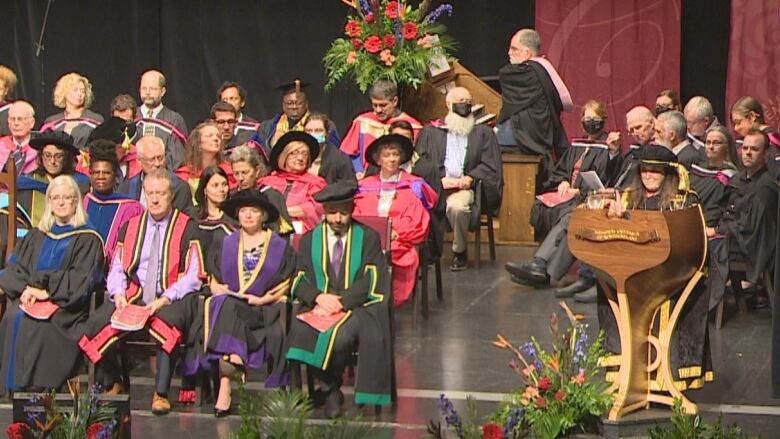 A group of academics in their regalia sit on chairs on a stage, while one speaks into microphone on a dais, and graduating students watch in the audience.