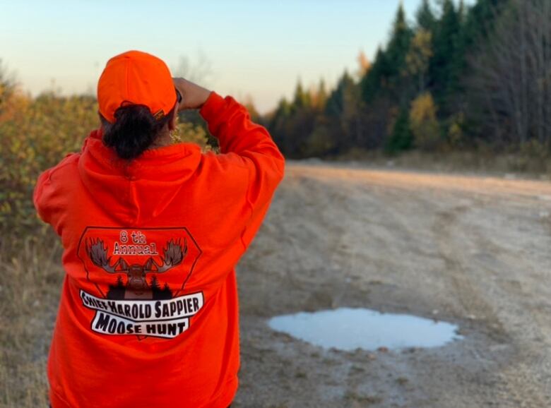 A woman in an orange sweater and hat stands with her back turned to the camera. She's pointing binoculars to the nearby woods.