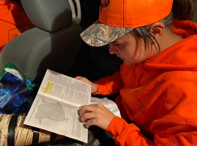 A young woman in an orange hat and baseball cap sits in a car reading a book.