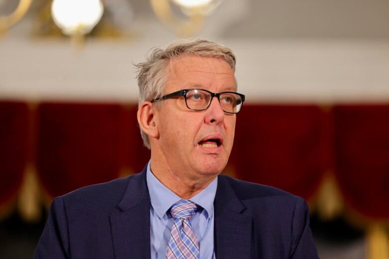 A man in a blue suit, with a blue shirt and plaid tie with glasses speaks.