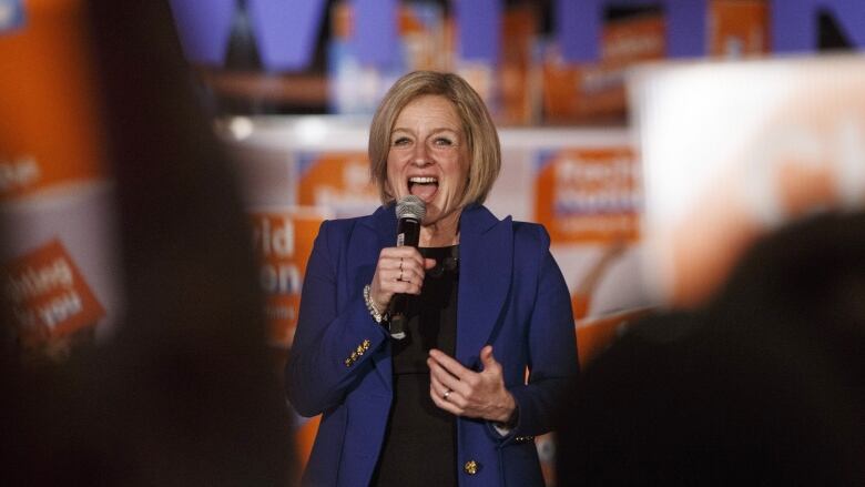 Rachel Notley stands centre-frame holding a microphone surrounded by blurred NDP signs and shadows of people in the foreground.