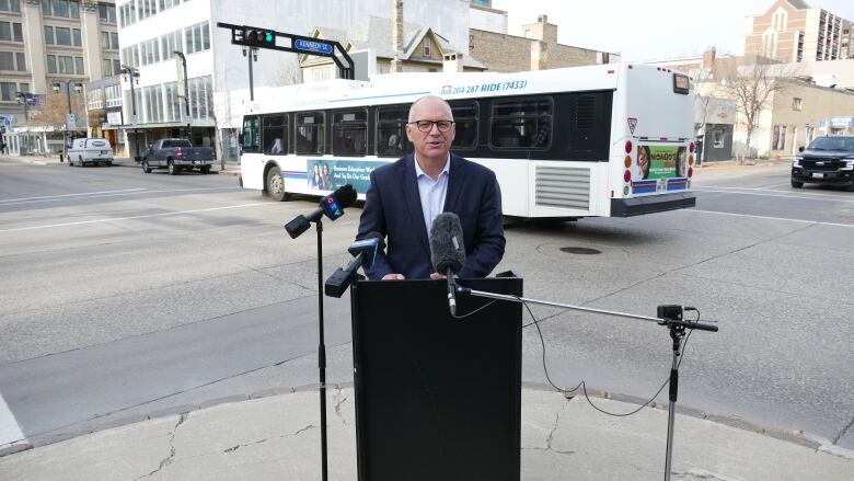 A man wearing a suit is speaking at a podium on a street corner with a bus driving past in the background.