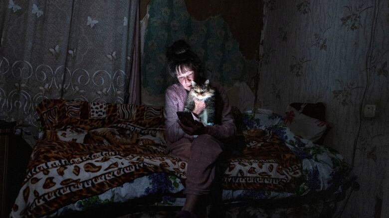 A woman sits on a bed in a dark room in her pajamas snuggling a cat, lit only by the light of her phone. 