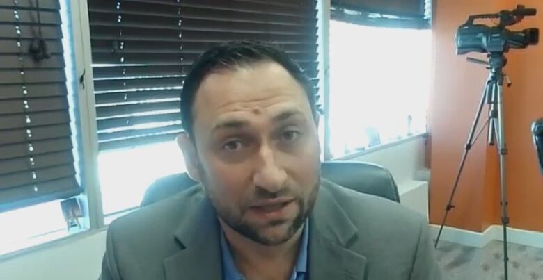 A man in a blazer speaks into a camera from an office with a window behind him.