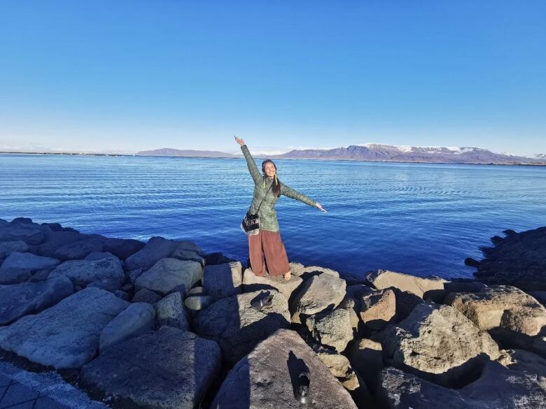 A woman poses on rocks by an expanse of water with mountains on the horizon.