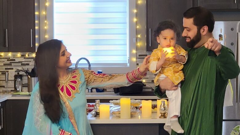 A man stands wearing green traditional Punjabi clothing, holding a one year old boy with curly hair, wearing a gold top and white pants. The baby is holding an LED candle with both hands as he looks into the camera. Left of them, a woman reaches out to the baby, wearing blue, pink and gold Punjabi dress smiling. They're in a kitchen with dark cupboards and light countertops, with candles and lights decorating the room.