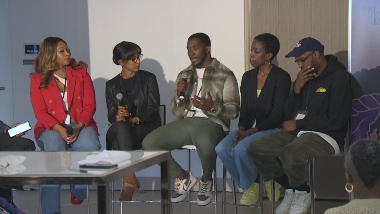 Five people sit side by side in chairs, holding microphones.