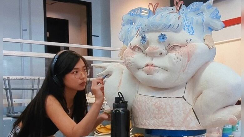 Photo of the artist Sami Tsang painting a large ceramic form of a plump cartoonish toddler with blue pigtails. She works in a white-walled artist studio. The artwork rests on a surface, and appears to be two or three feet taller than the artist.