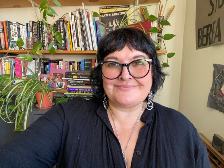 A selfie photo of a person wearing a black dress shirt, black-rimmed glasses and silver jewellery, in front of a wall with books and plants on shelves.