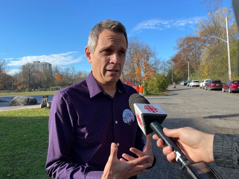 A man in a purple shirt speaks to a reporter on a sunny day.