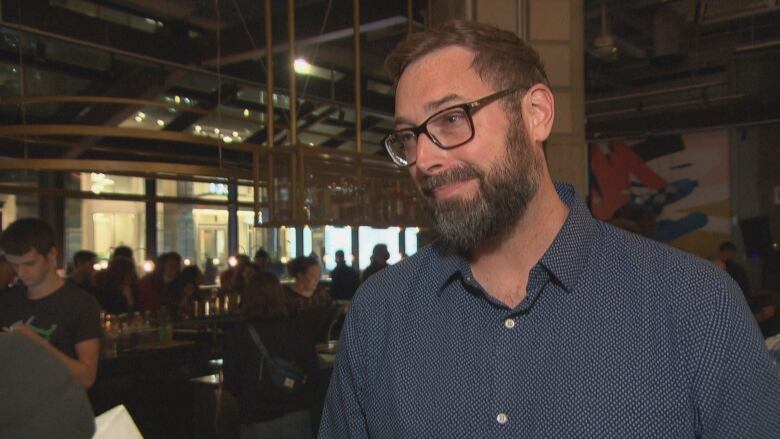 A man with a beard stands in a room while people eat and drink in the background.