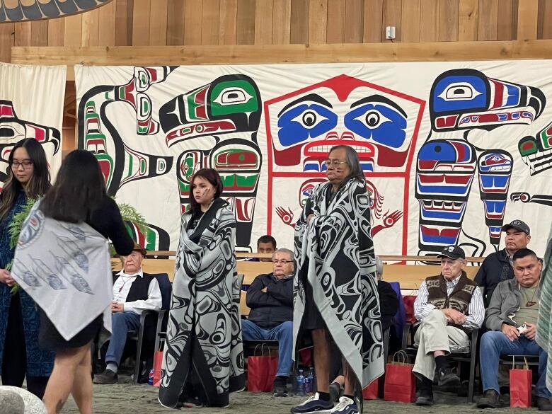 An Indigenous man and teenage girl wearing traditional clothing stand among a small crowd in front of Indigenous artwork.