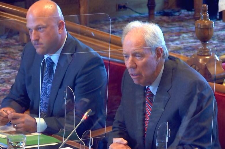 A man wearing a business suit speaks at a desk in a legislature. Another man in a business suit sits to his right. Plexiglas shields line the desk as a social distancing measure. 