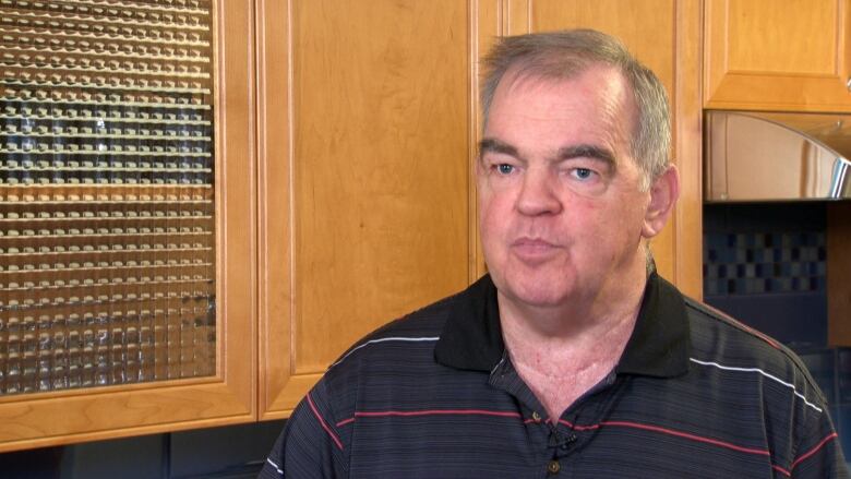 A man with thinning grey hair, in a polo shirt, stands in front of wooden kitchen cabinets. 